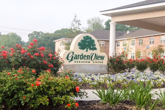 Entrance sign for Garden View Assisted Living surrounded by blooming red and white flowers, with a brick building and covered entrance in the background.