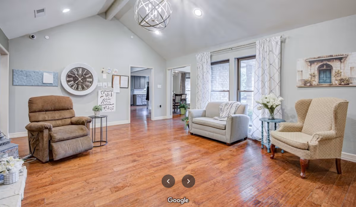 A bright living room with wooden flooring, featuring a brown recliner chair, a small round side table with a plant, a light gray loveseat with a throw blanket, and a beige upholstered armchair next to a small round table with a vase of white flowers. The walls are light gray with a large clock, bulletin board, and framed artwork. Large windows with white patterned curtains let in natural light.