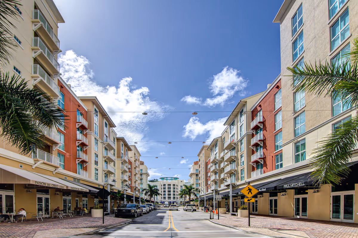 Pedestrian-friendly street lined with multi-story residential buildings with ground-floor shops and palm trees under a blue sky.