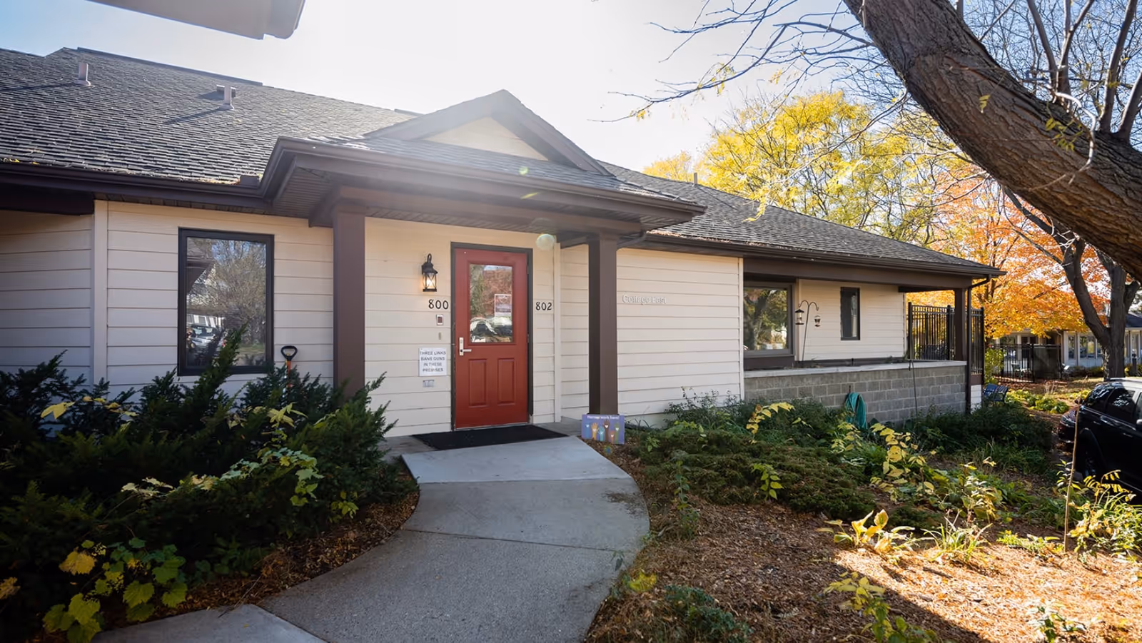 Front entrance of a single-story senior living facility with red door and landscaped walkway.