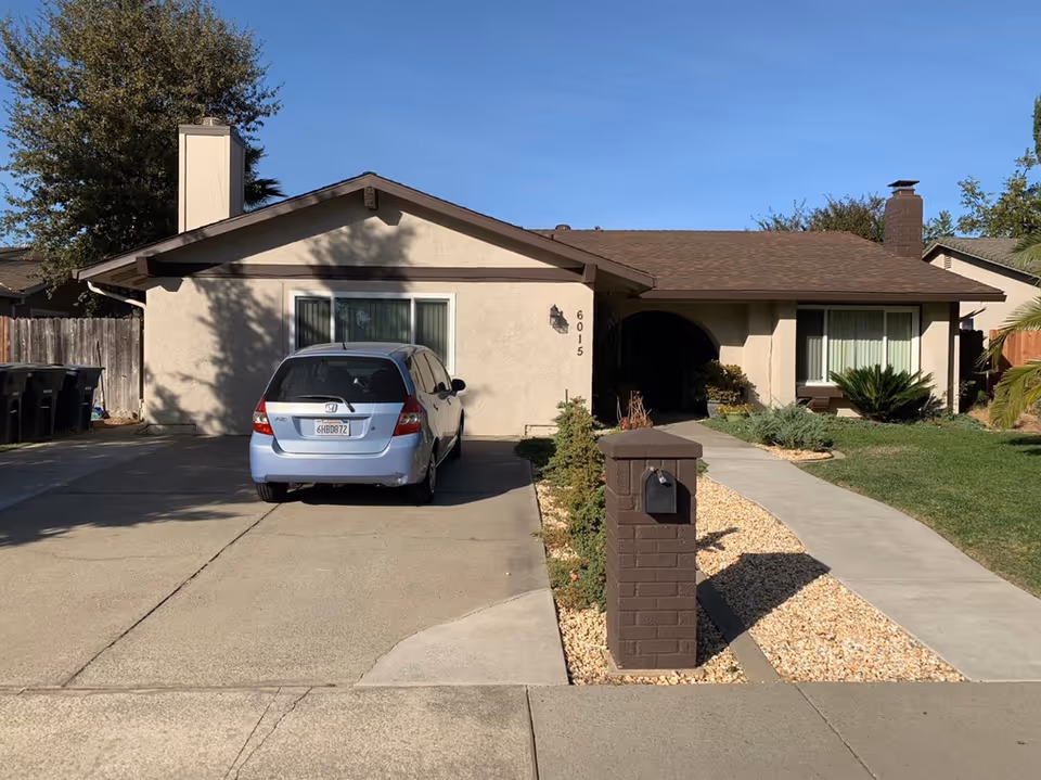 Single-story home front with a driveway and parked car, mailbox, walkway and small lawn under a clear sky.