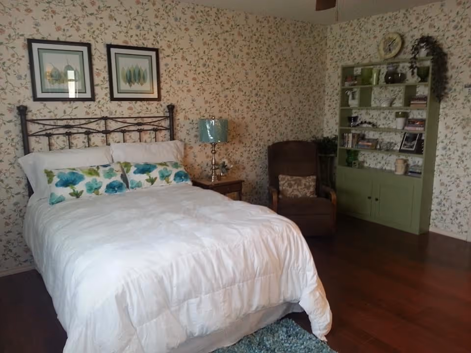 Cozy bedroom with white comforter, floral wallpaper, metal bed frame, and a green bookshelf.