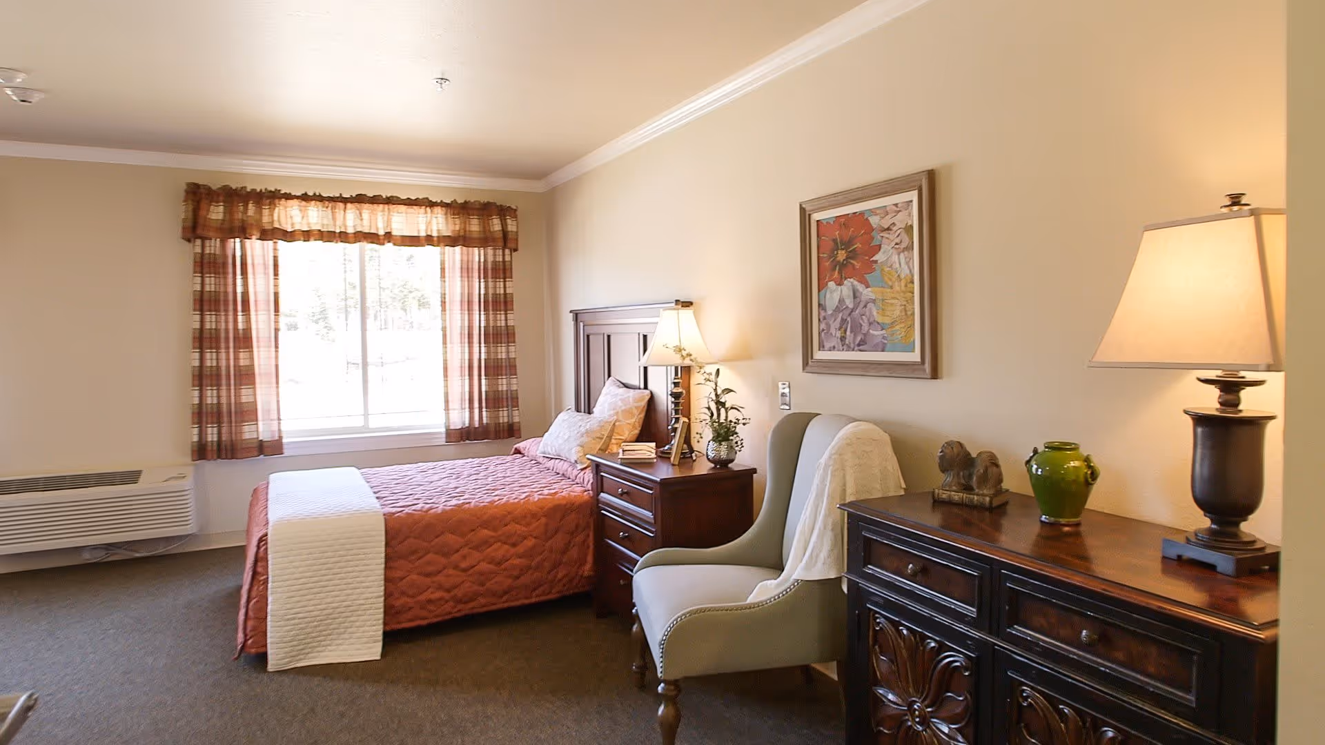 A cozy bedroom in an assisted living facility featuring a single bed with a red quilt and white blanket, a wooden nightstand with a lamp and decorative items, a comfortable armchair with a white throw draped over it, a wooden dresser with a lamp, green vase, and small sculpture, and a window with plaid curtains allowing natural light to fill the room.