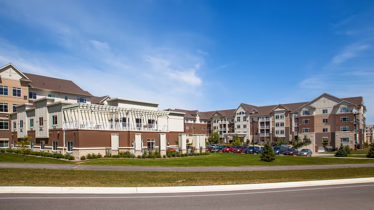 Exterior view of a multi-story senior living facility with a well-maintained lawn, parking lot with cars, and clear blue sky.