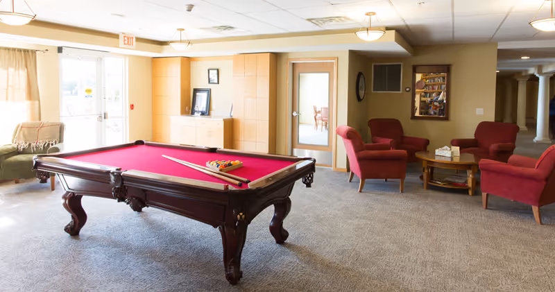A bright communal lounge with a red-felt pool table and grouped red armchairs around a coffee table.