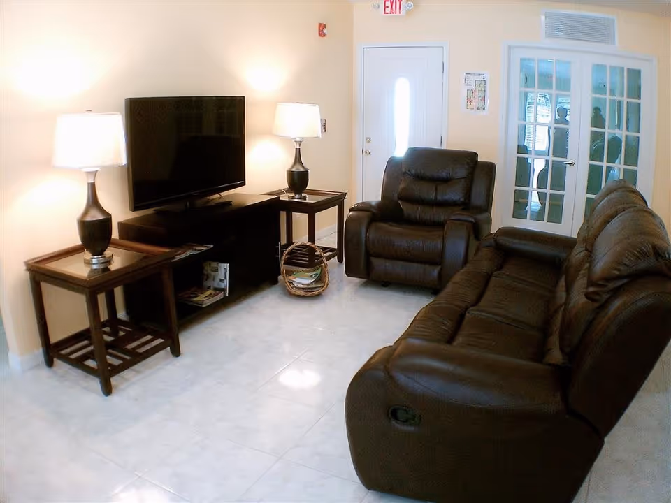 Bright living room with brown leather recliner and sofa facing a flat-screen TV on a stand, two table lamps, and glass-paneled doors.