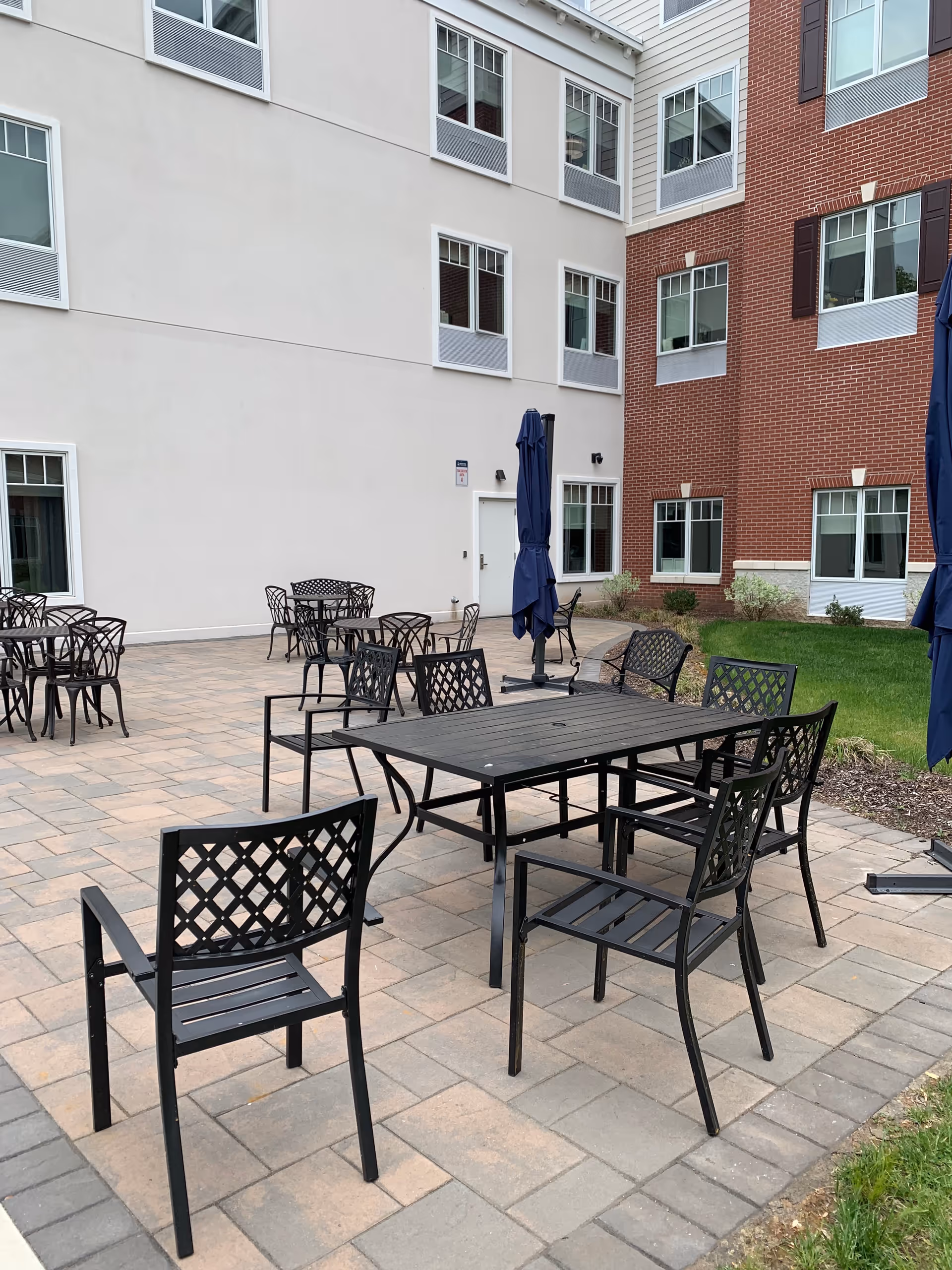 Outdoor patio with metal tables, chairs, and folded umbrellas near a multi-story building.