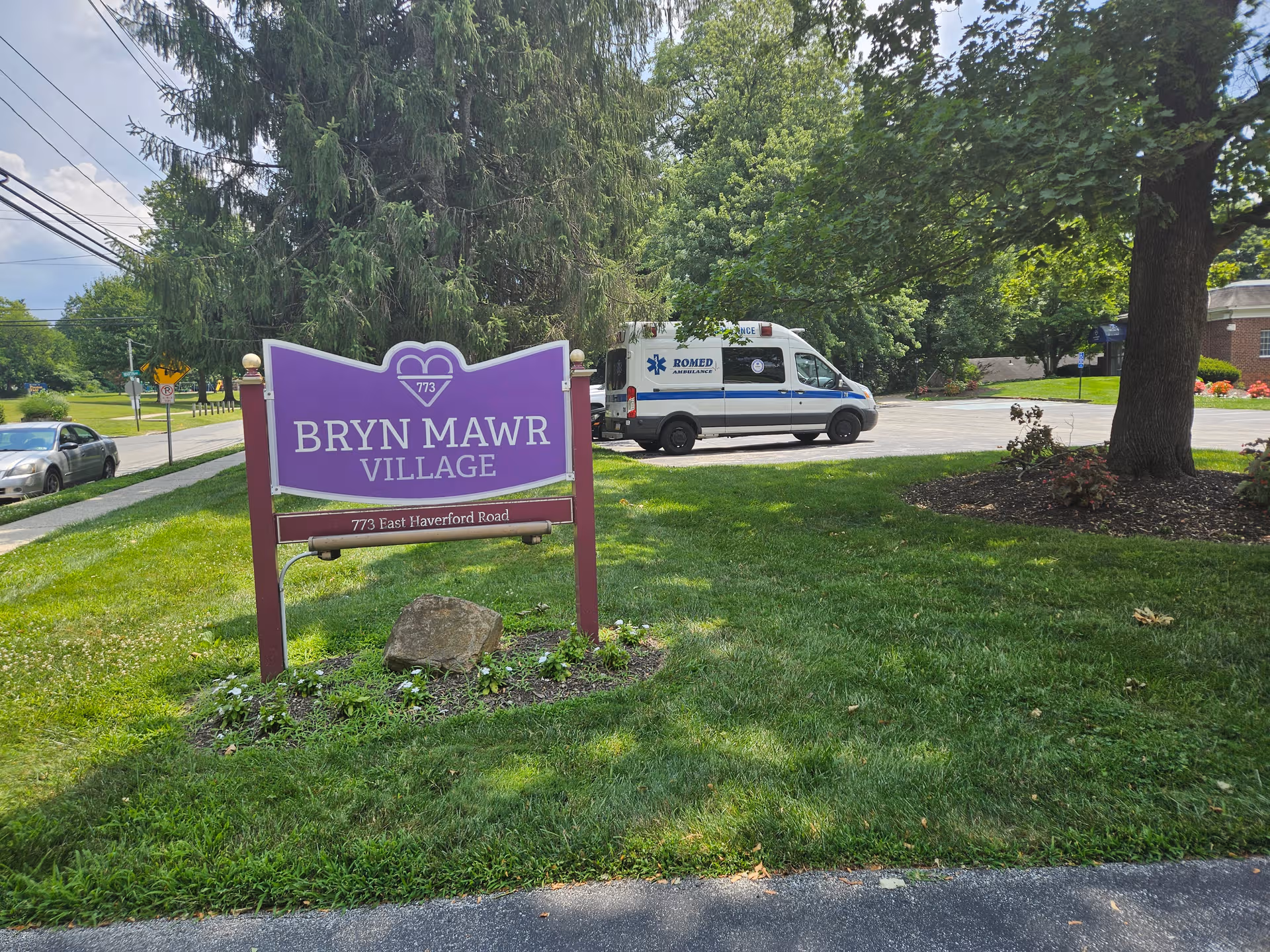Purple Bryn Mawr Village sign sits on a grassy lawn with an ambulance nearby.