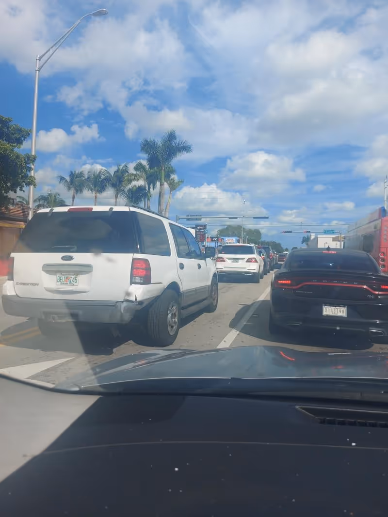Street view from inside a car; white SUV, traffic, palm trees, blue sky.