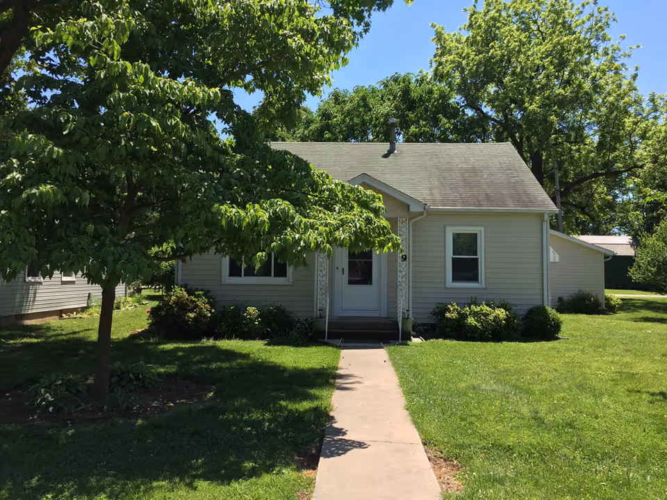 Front exterior of a small single-story house with a walkway, lawn, and trees.