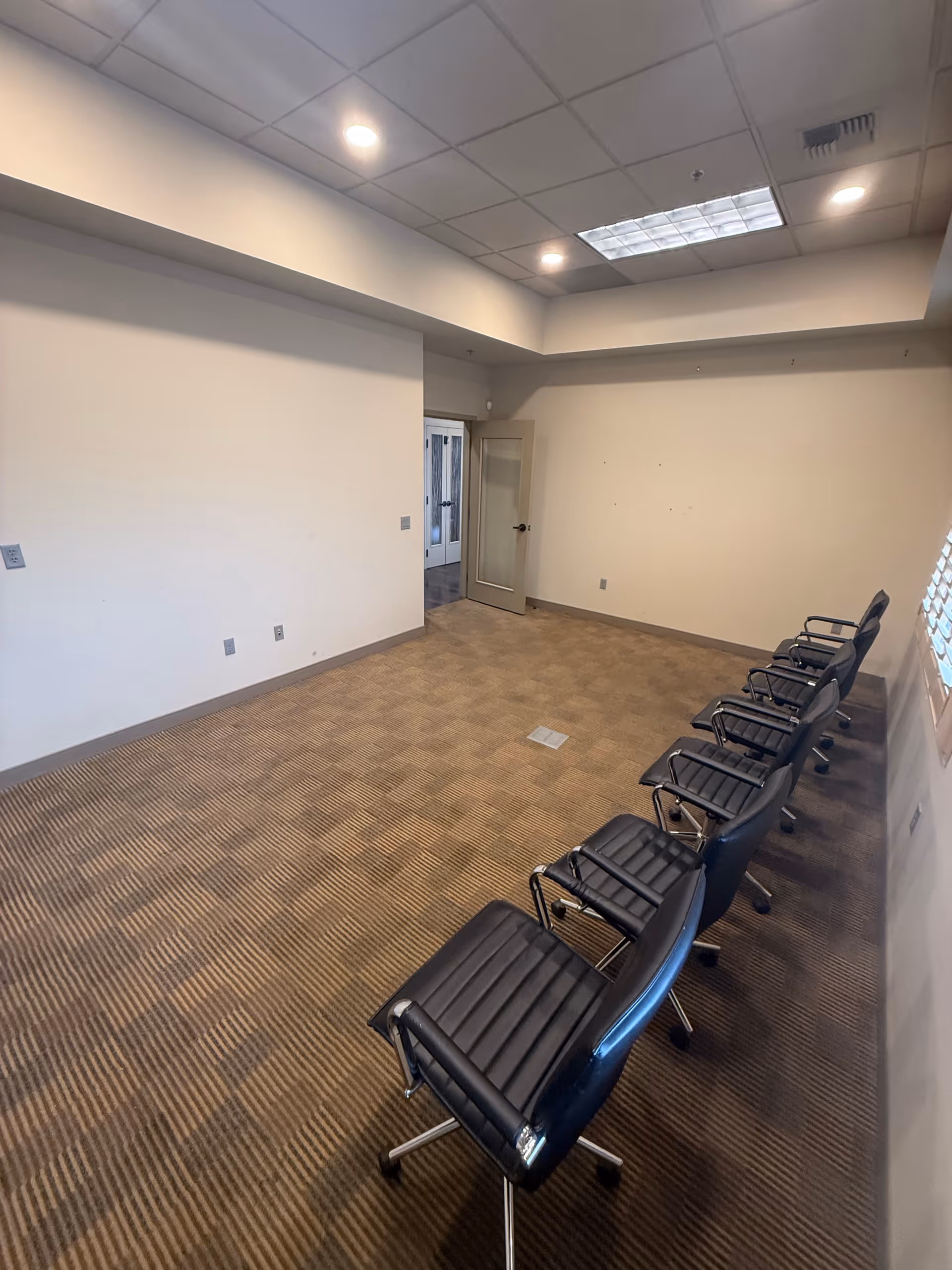 A sparsely furnished conference room with black chairs along the wall and open doorway.