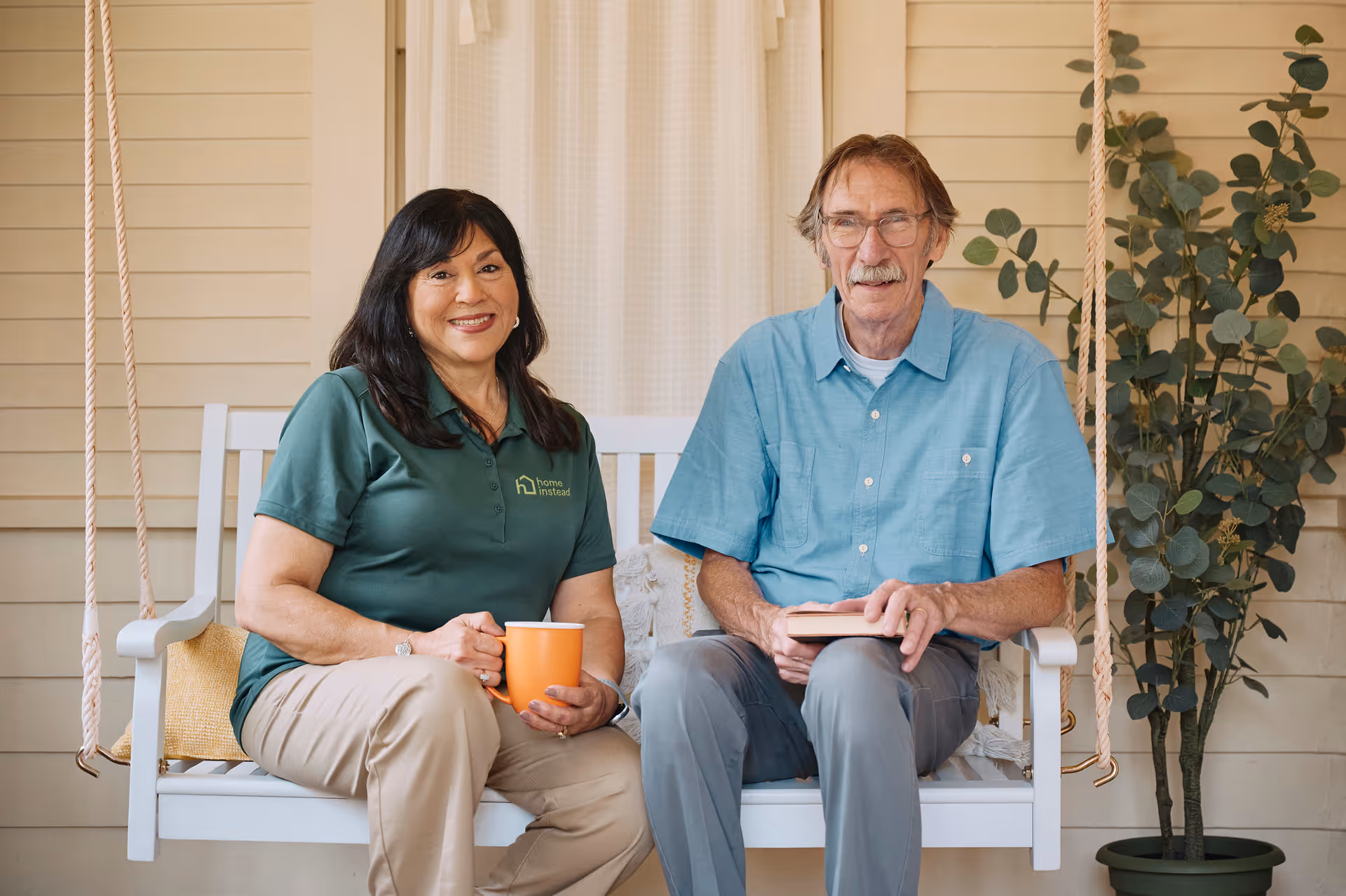 Two smiling adults sit on a white outdoor porch swing, holding a mug and a book.