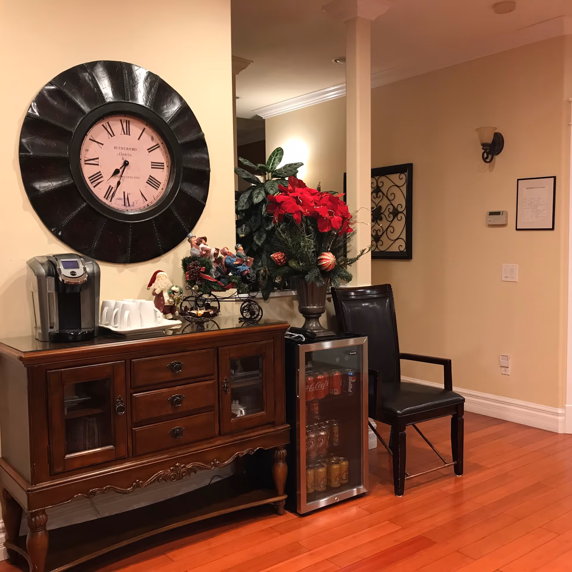 Warm lobby area with a large wall clock, wooden sideboard, beverage refrigerator, and holiday decor.