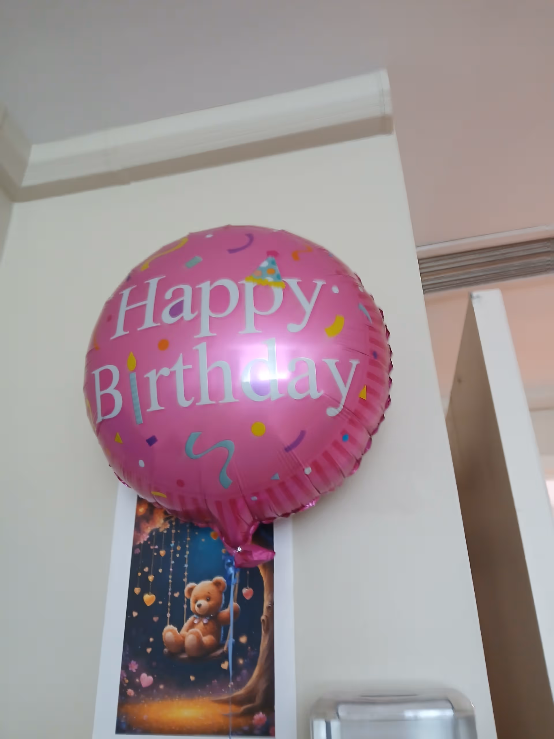 Pink Happy Birthday balloon indoors near a teddy bear poster.
