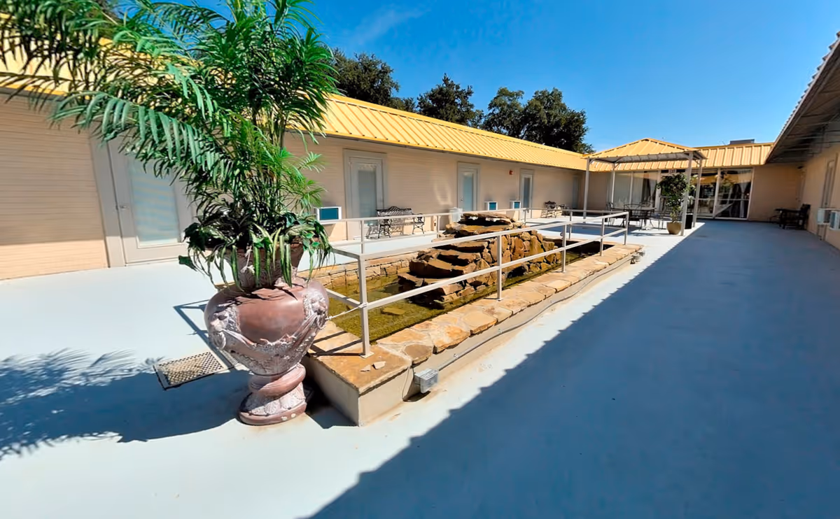 Sunny enclosed courtyard with a central rock water feature, potted plants, and surrounding walkways and doors.
