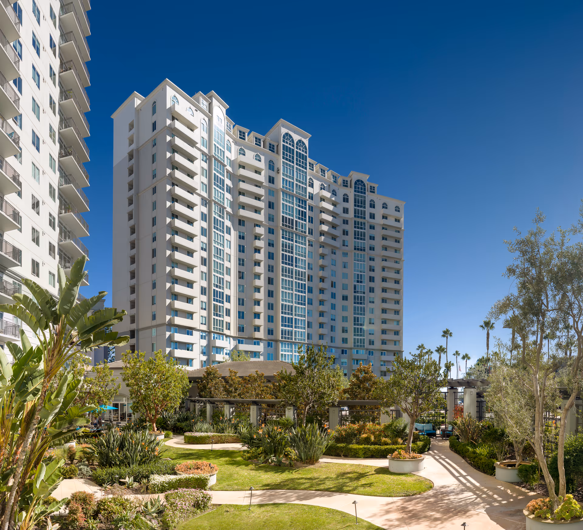 Tall white senior living building with balconies and landscaped courtyard under blue sky.