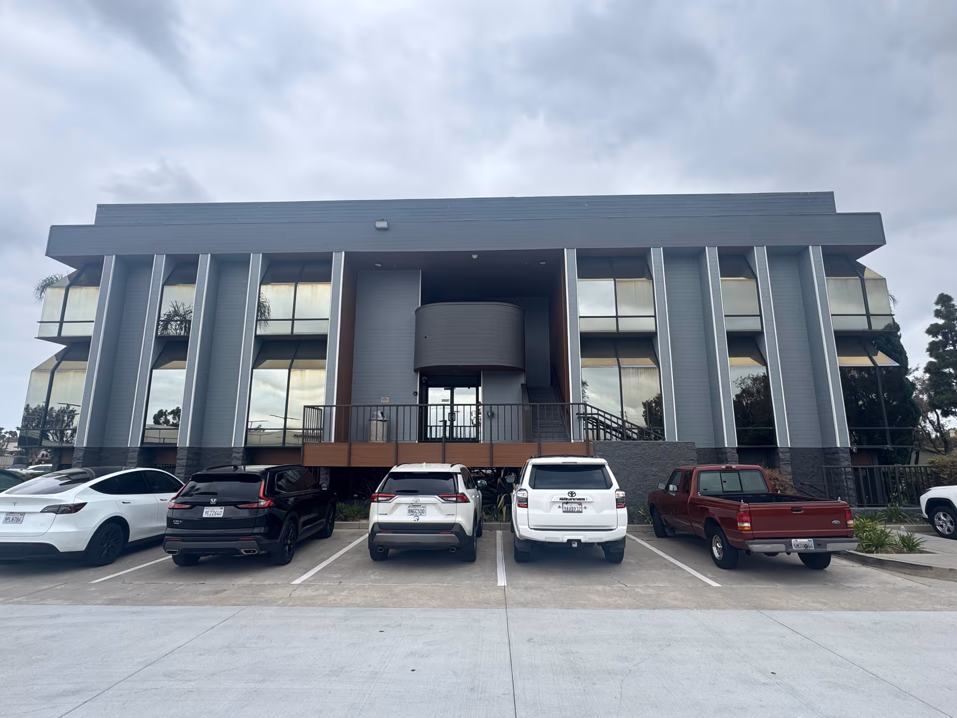 Modern gray building with large glass windows and several parked cars in front.