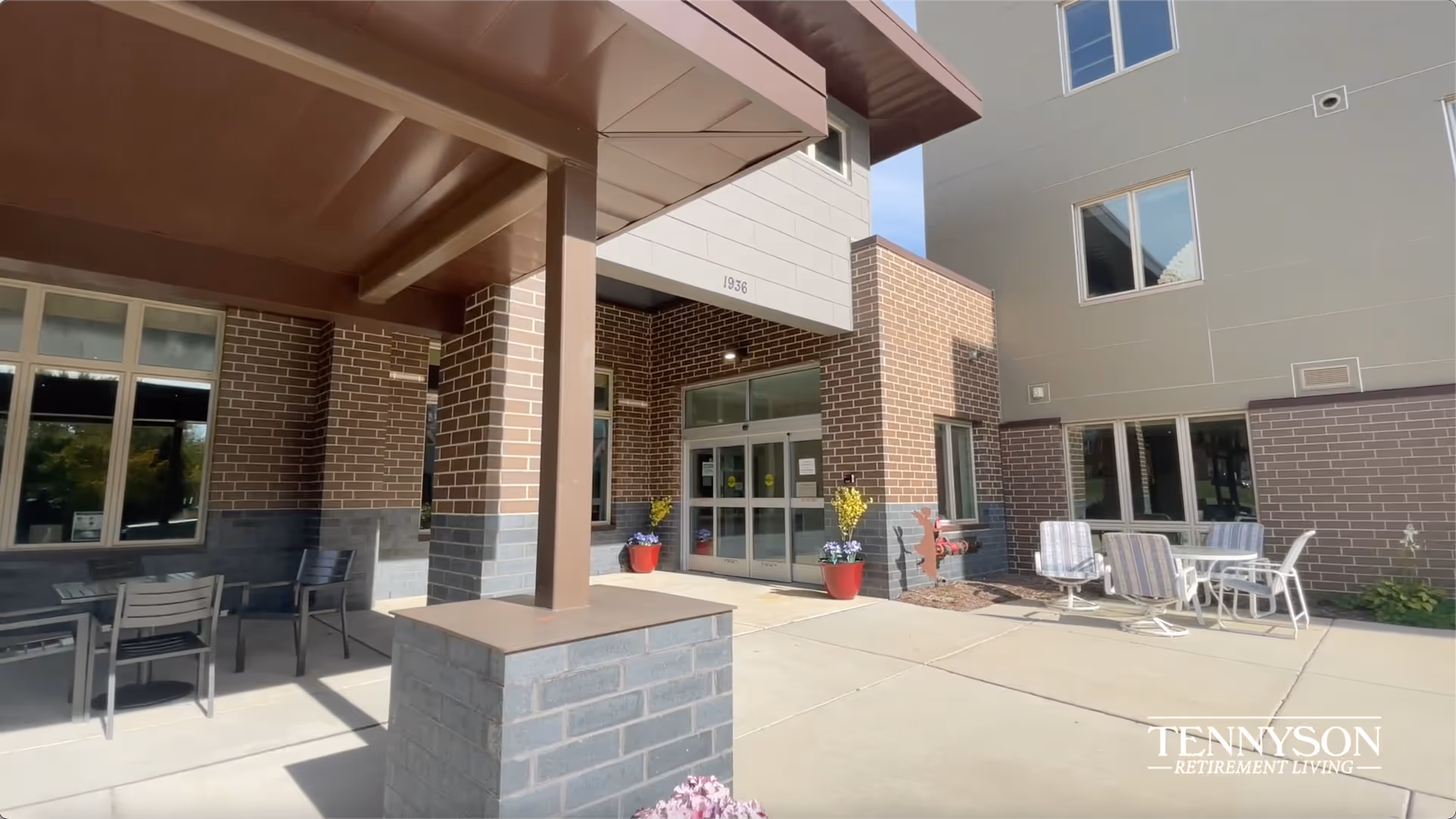 Exterior entrance of The Sonnet at Tennyson Assisted Living & Memory Care facility showing a covered walkway with brick pillars, glass double doors, potted plants, and outdoor seating with tables and chairs on a sunny day.