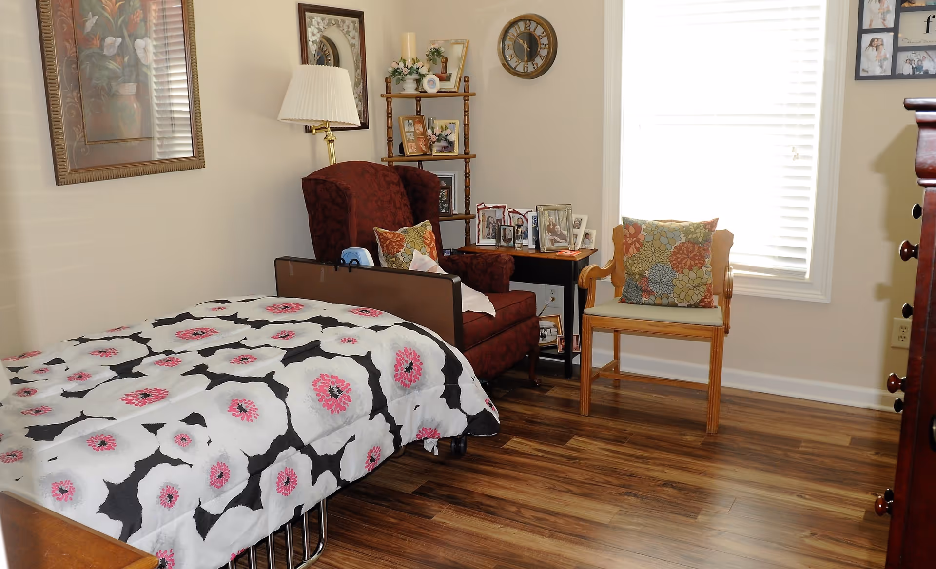 A cozy bedroom with a single bed covered in a white, black, and pink floral patterned blanket. Next to the bed is a burgundy armchair with a decorative pillow, a small side table with framed photos, and a wooden chair with a colorful floral cushion. The room has wooden flooring, a window with white blinds, a wall clock, and framed pictures on the walls.