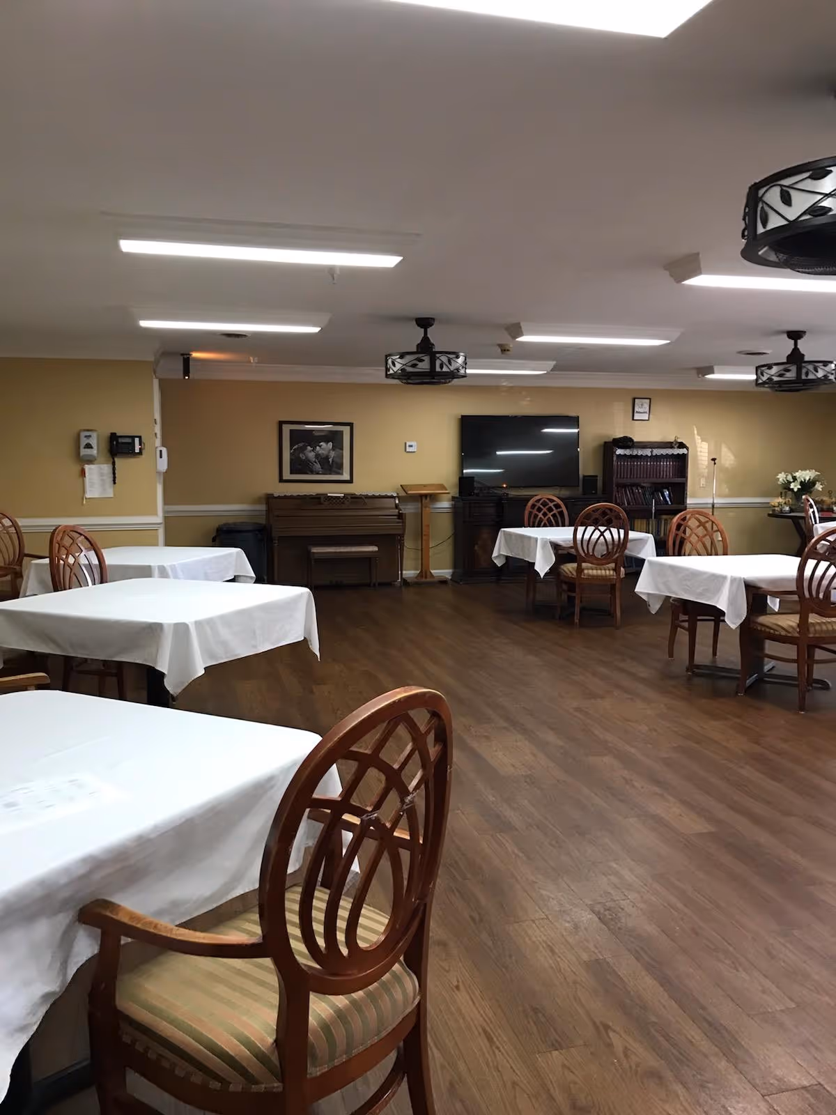 Spacious dining/activity room with tables covered in white tablecloths, wooden chairs, a TV and piano along the far wall.