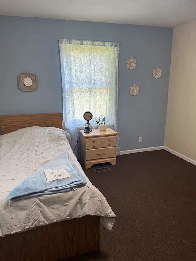 Bedroom with bed, nightstand, lace-curtained window, and soft blue walls.