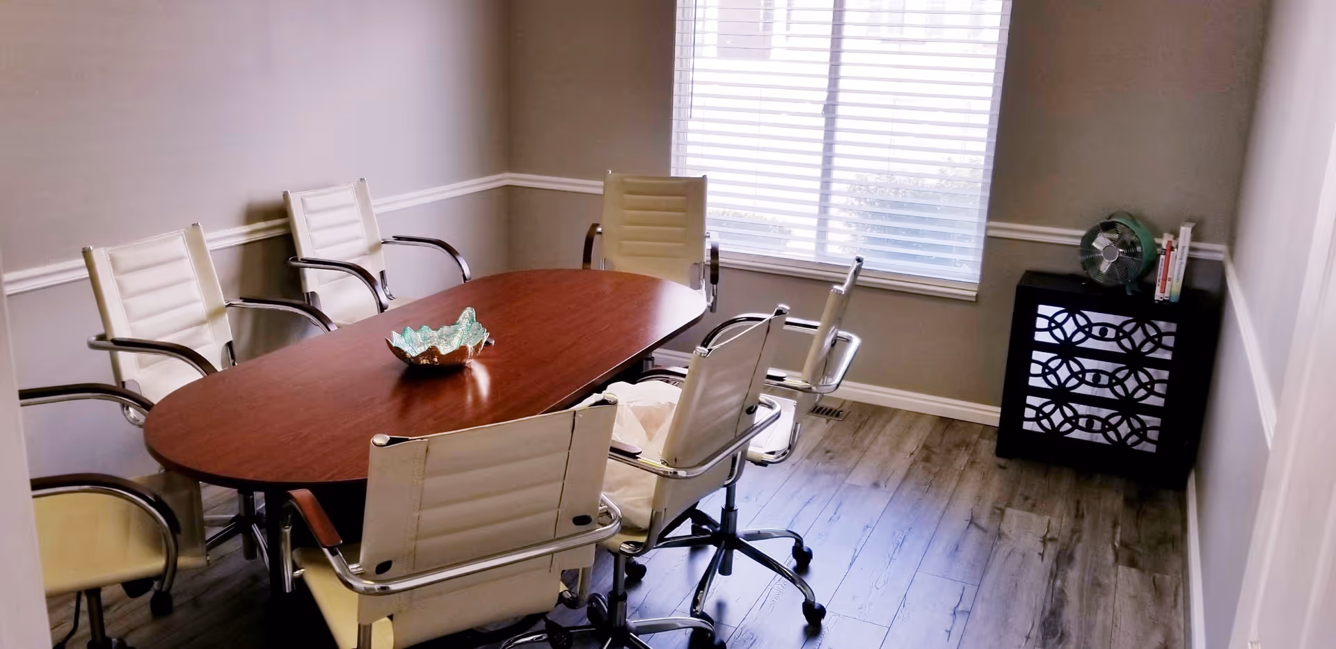 Oval wooden conference table surrounded by white office chairs in a bright meeting room.