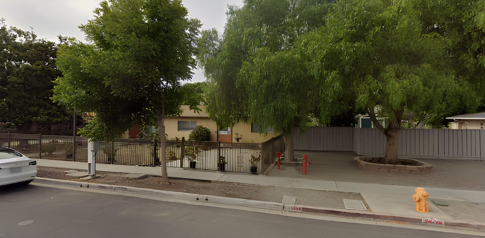 Street view showing the exterior of a single-story building partially obscured by large green trees. There is a sidewalk, a white car parked on the street, a fire hydrant, and a metal fence surrounding the property.