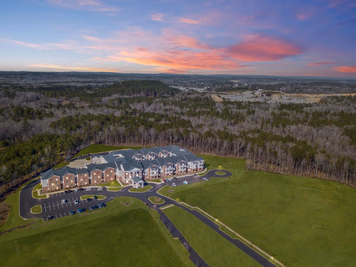 Aerial view of Legacy Reserve at Old Town, a large senior living facility surrounded by green lawns and dense forest under a colorful sunset sky.