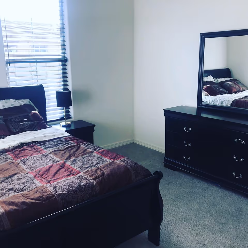 A dark wooden bedroom featuring a bed, dresser with mirror, nightstand, and window blinds.