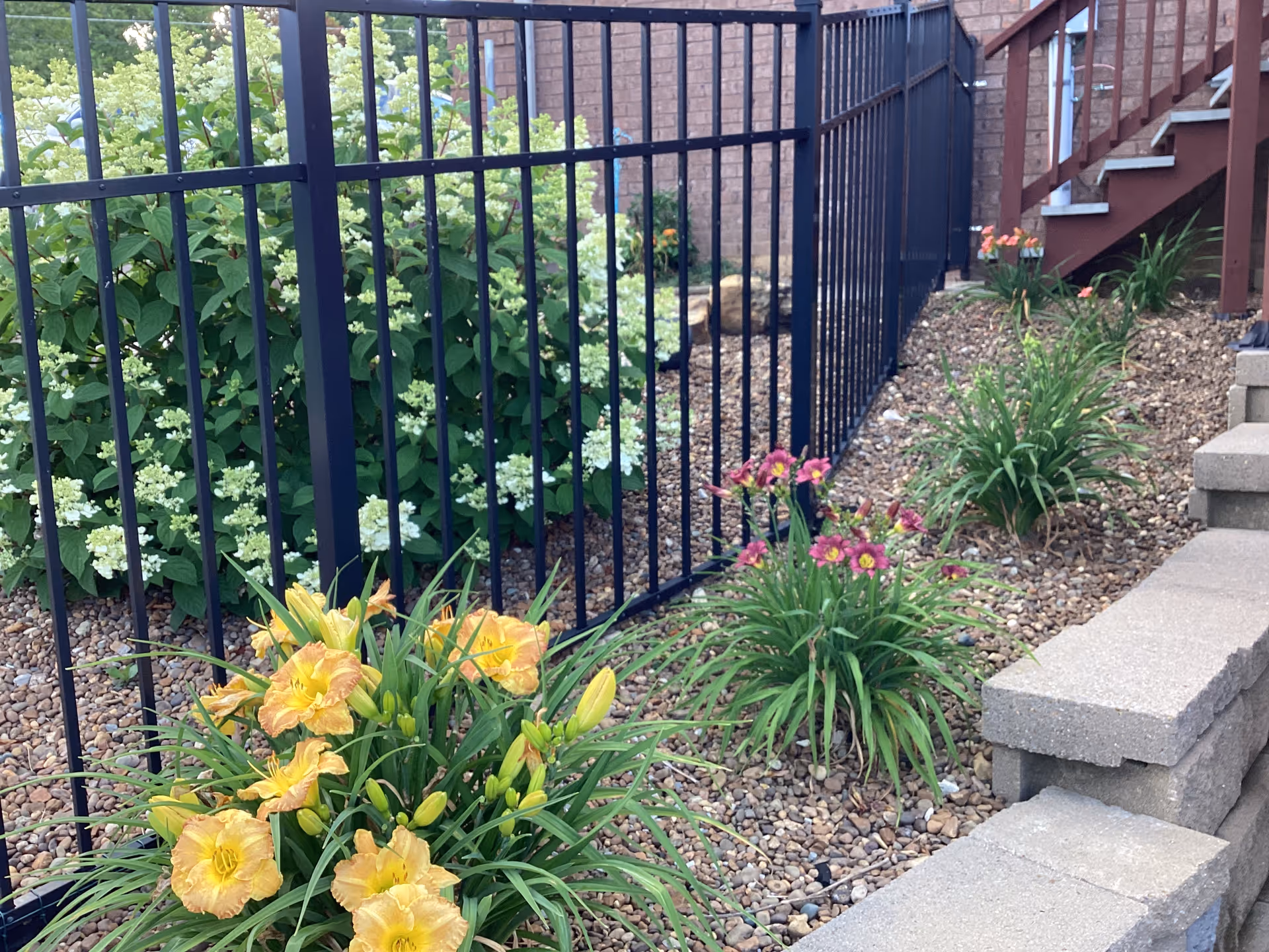 Exterior view of a fenced, landscaped walkway with flowering plants beside a brick building and stairs.