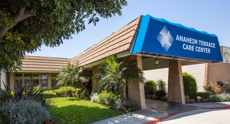 Front entrance of Anaheim Terrace Care Center with a blue awning over a covered driveway, brick pillars, and landscaped greenery.