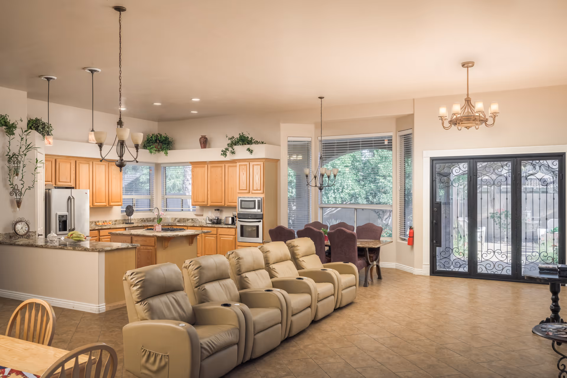 Open-plan interior common area with a kitchen, dining table, and a row of beige recliner chairs near decorative double doors.