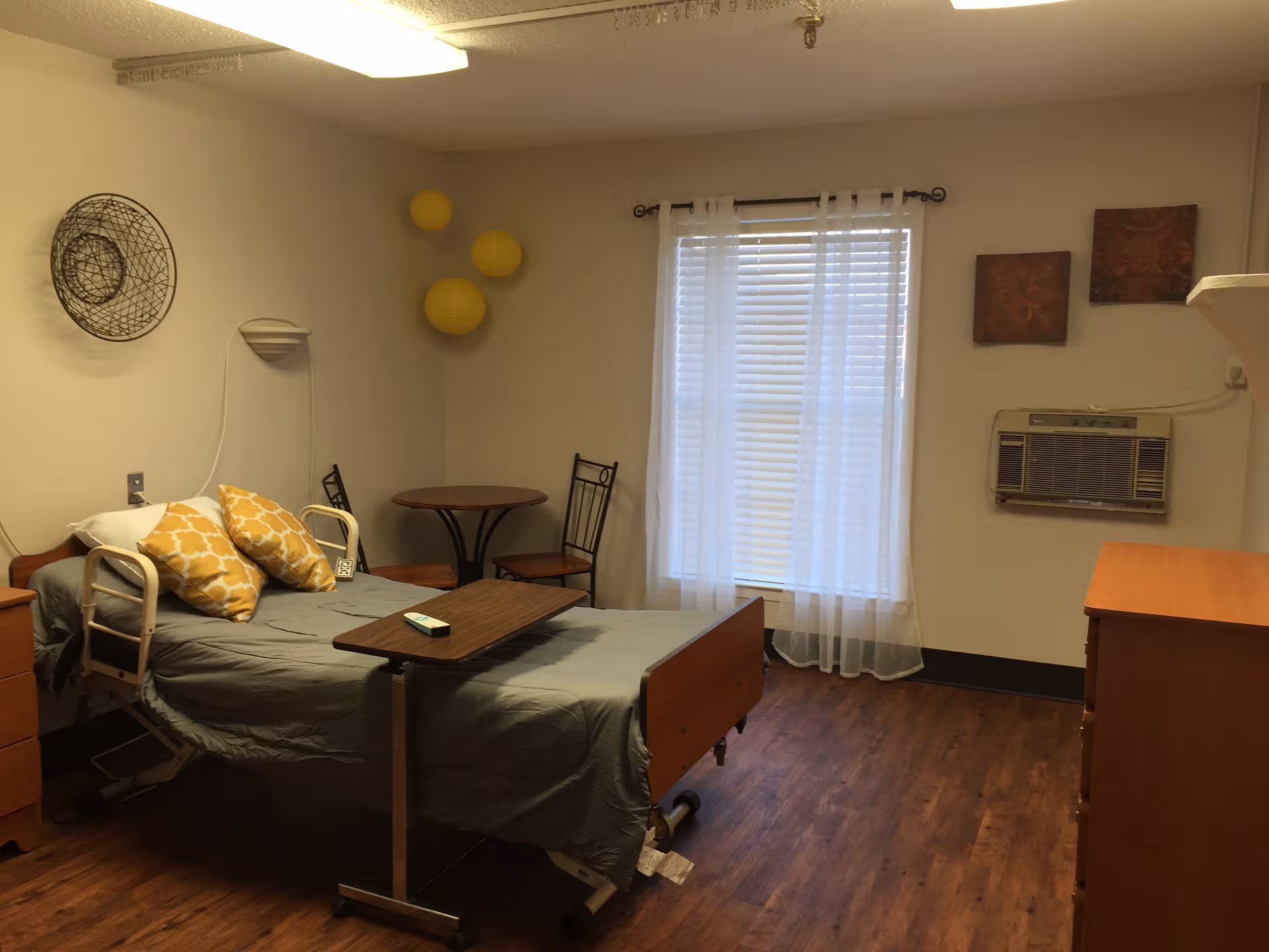 A hospital-style patient room with a bed, dresser, window, and seating area.