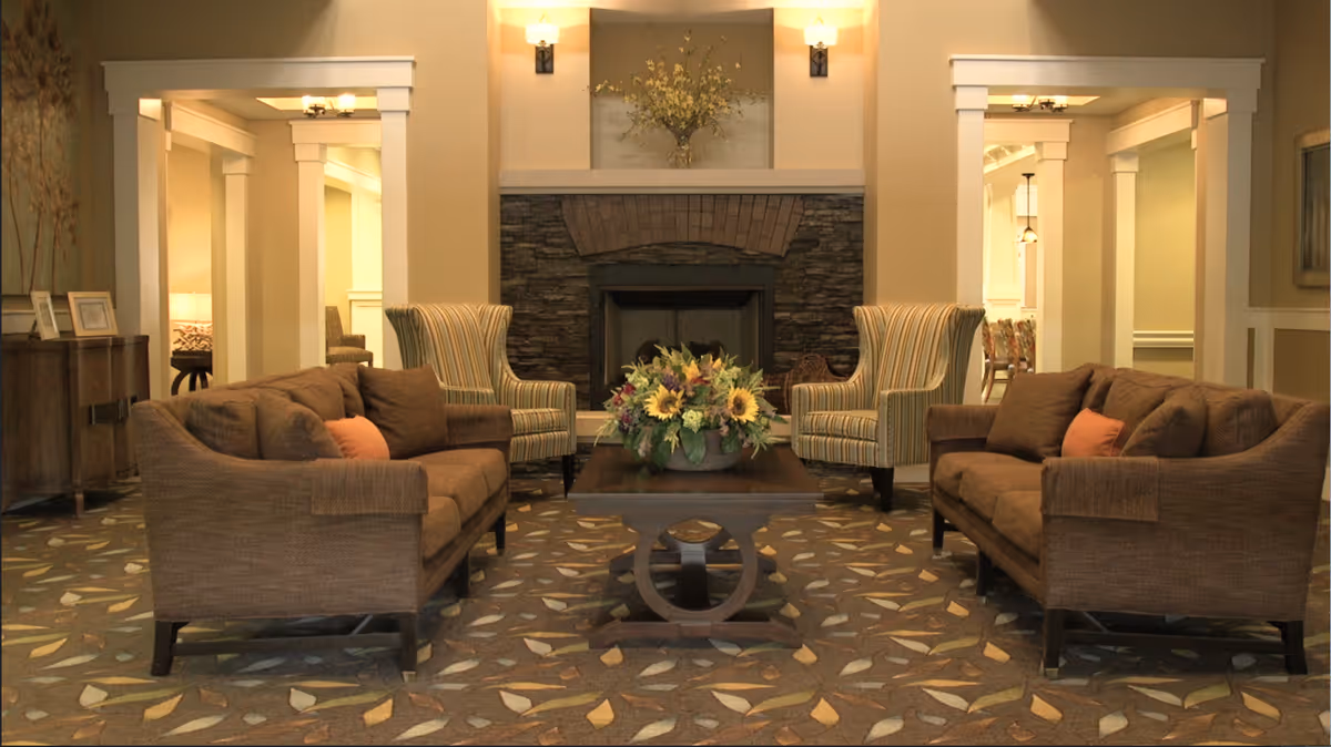 Cozy senior living lounge with brown sofas, striped chairs, fireplace, and floral centerpiece.