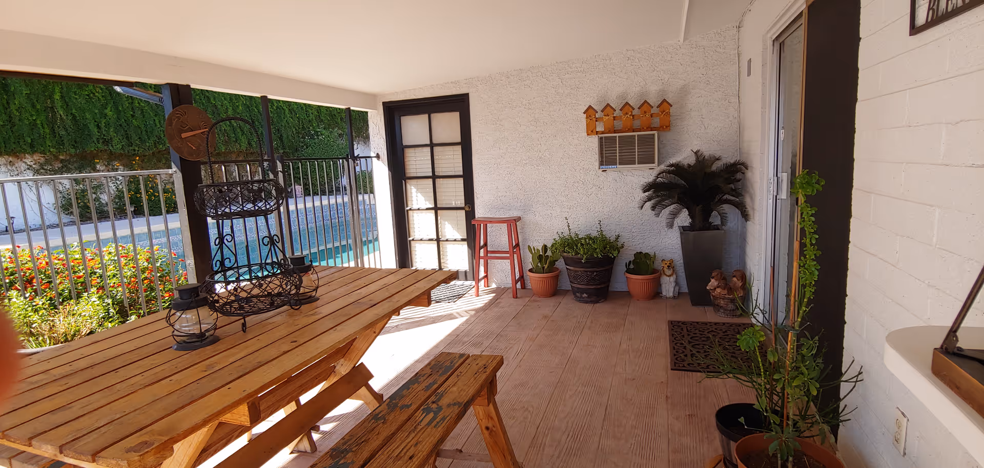 Covered outdoor patio area with a wooden picnic table and bench, various potted plants, a small red stool, a wall-mounted air conditioner, and a door leading inside. The patio overlooks a swimming pool and garden area with greenery and flowers.