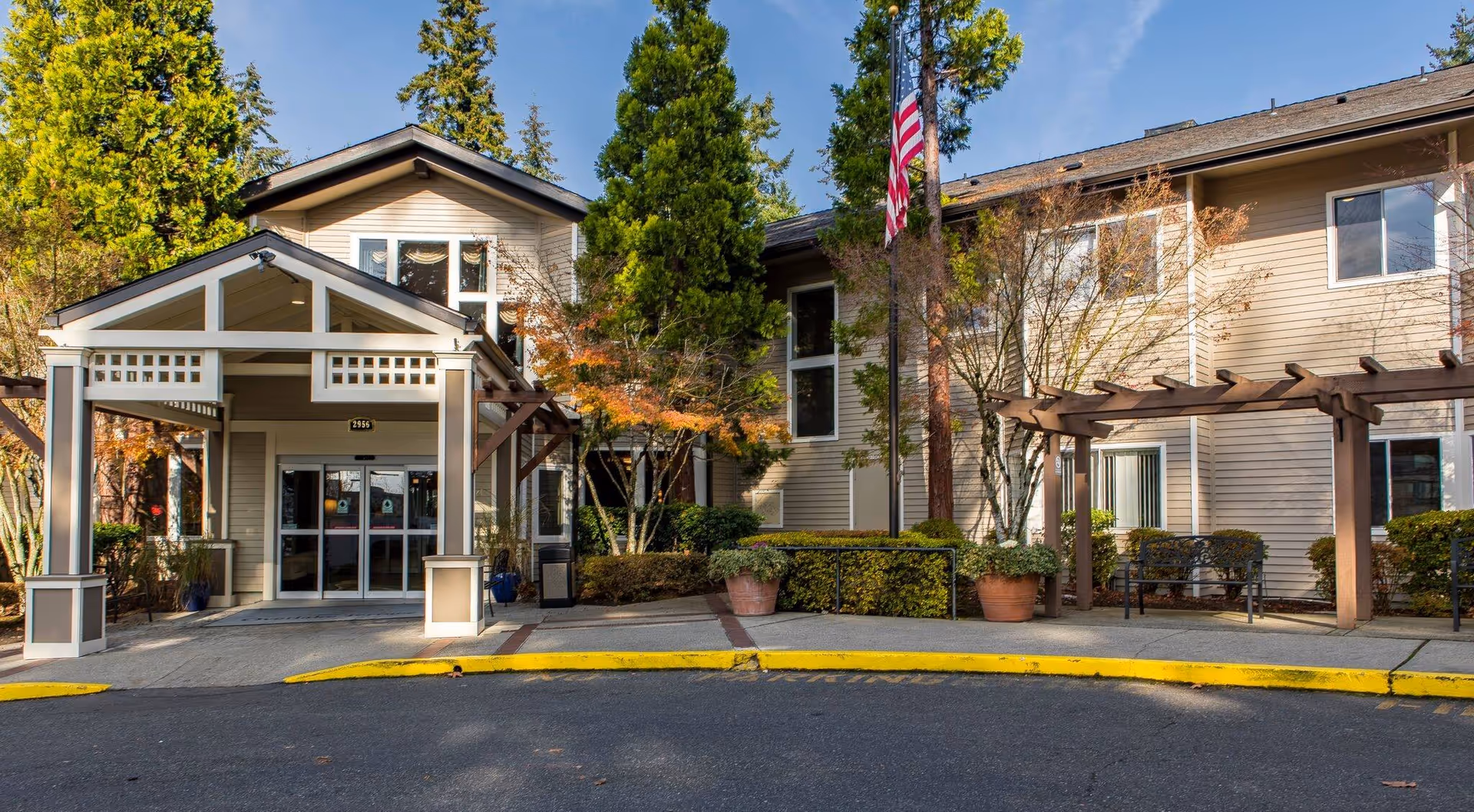 Front entrance of a senior living building with a covered porte-cochere, potted plants, a pergola, and an American flag.