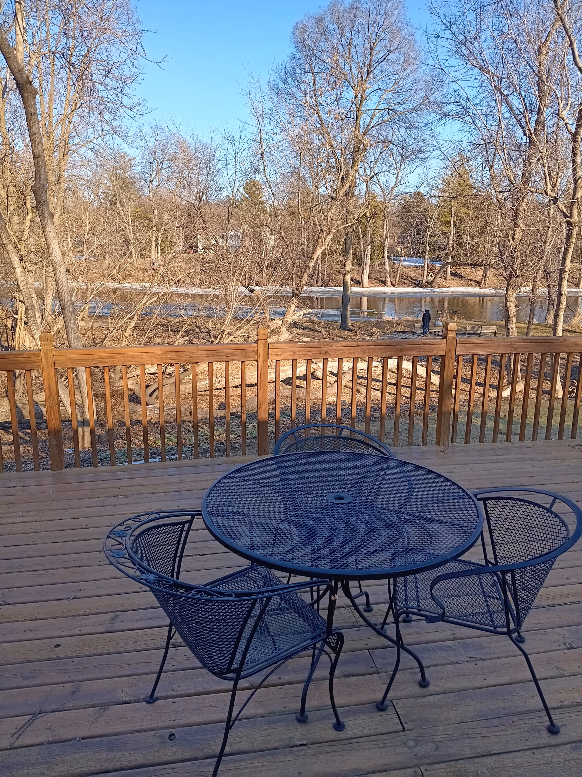 Outdoor wooden deck with blue metal table and chairs, overlooking trees and a water view.