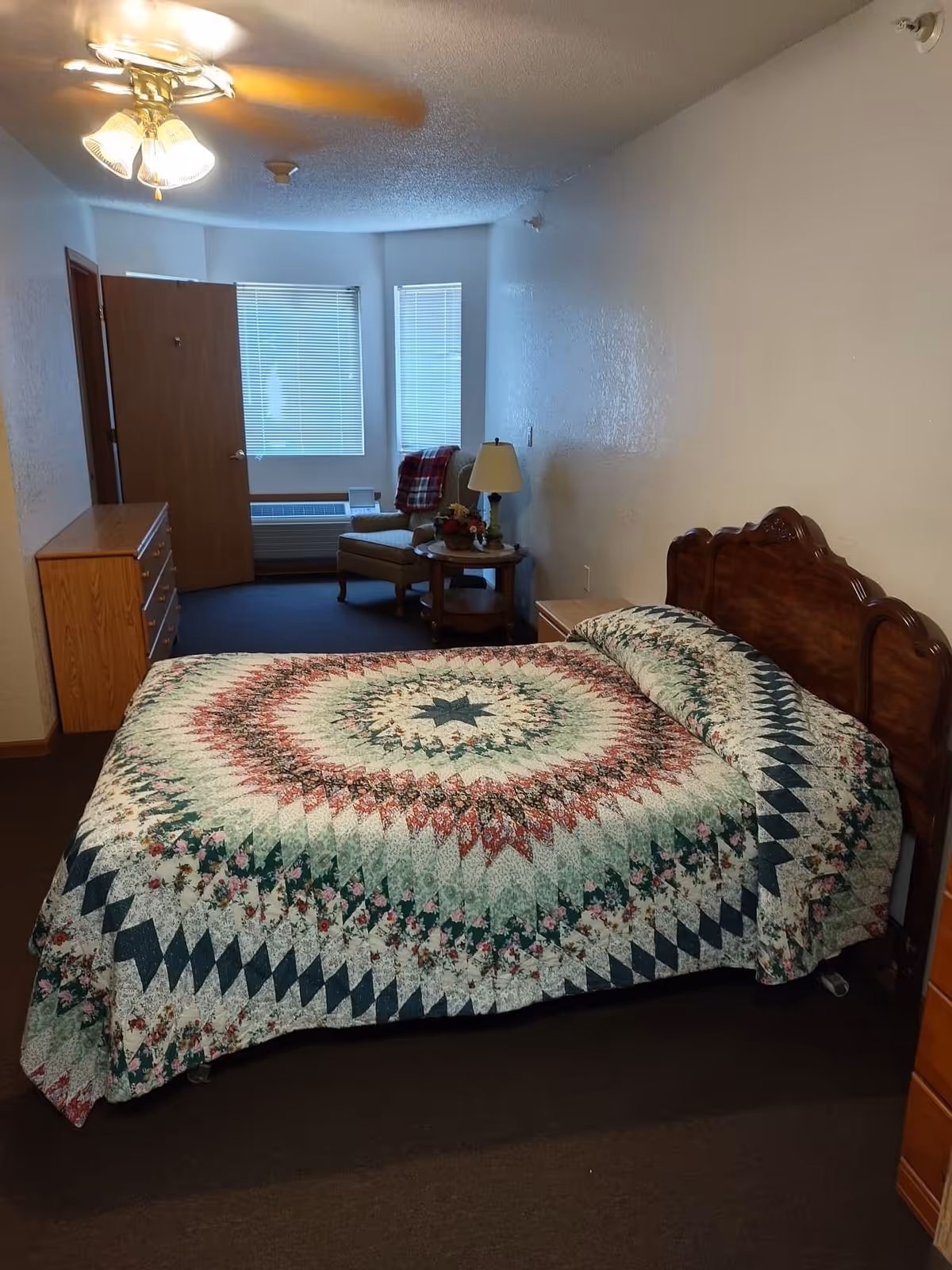A bedroom with a large bed covered in a colorful quilt featuring a circular pattern. The room has a wooden headboard, a wooden dresser on the left, and a small wooden nightstand with a lamp and a flower arrangement. There is a comfortable armchair with a red and black plaid blanket draped over it near two windows with blinds. The door to the room is open, and a ceiling fan with lights is visible above.