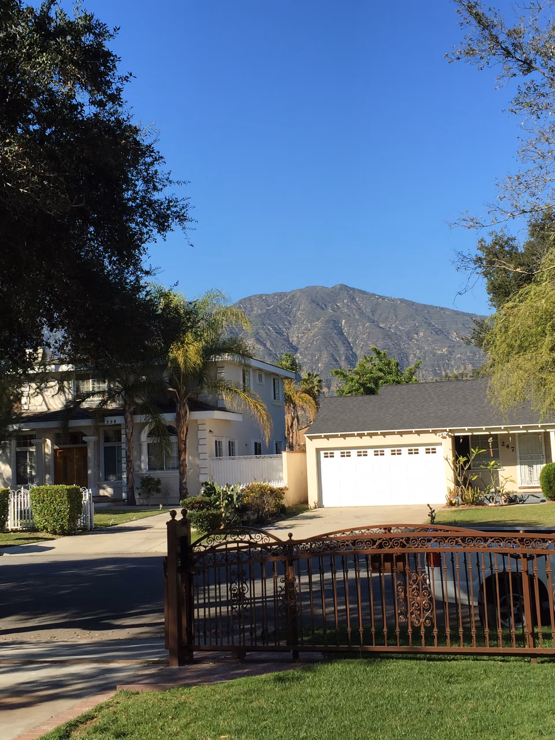 View of a residential street with two houses, one with a white garage door and the other a two-story house partially obscured by trees. There is a decorative metal gate in the foreground and a mountain range in the background under a clear blue sky.