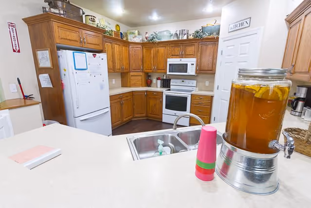 Communal kitchen with oak cabinets and white appliances, a sink in the foreground and a large glass beverage dispenser on the counter.
