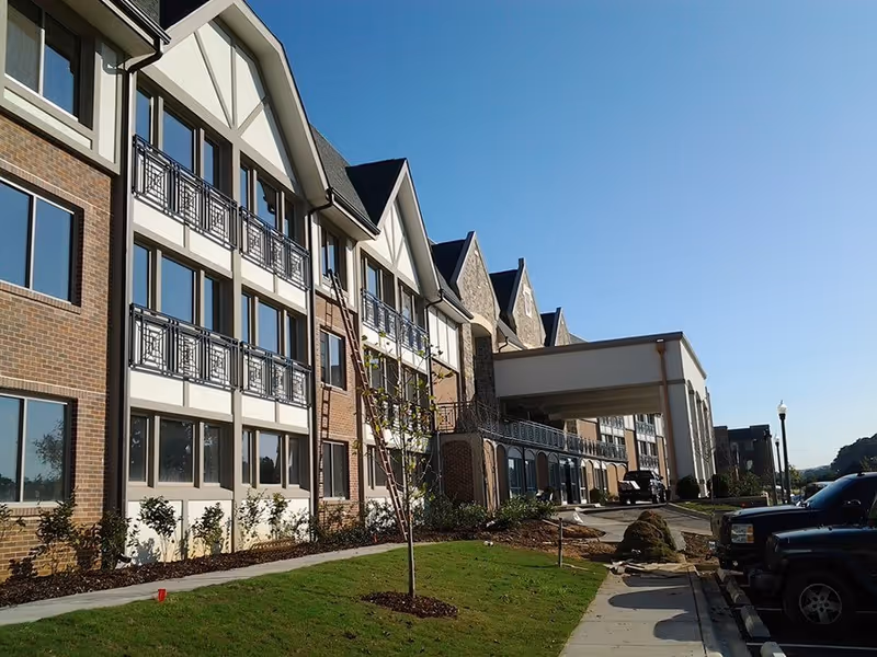 Exterior view of a multi-story senior living facility building with brick and white paneling, multiple windows, a covered entrance, a small landscaped lawn with young trees, and parked cars along the driveway under a clear blue sky.
