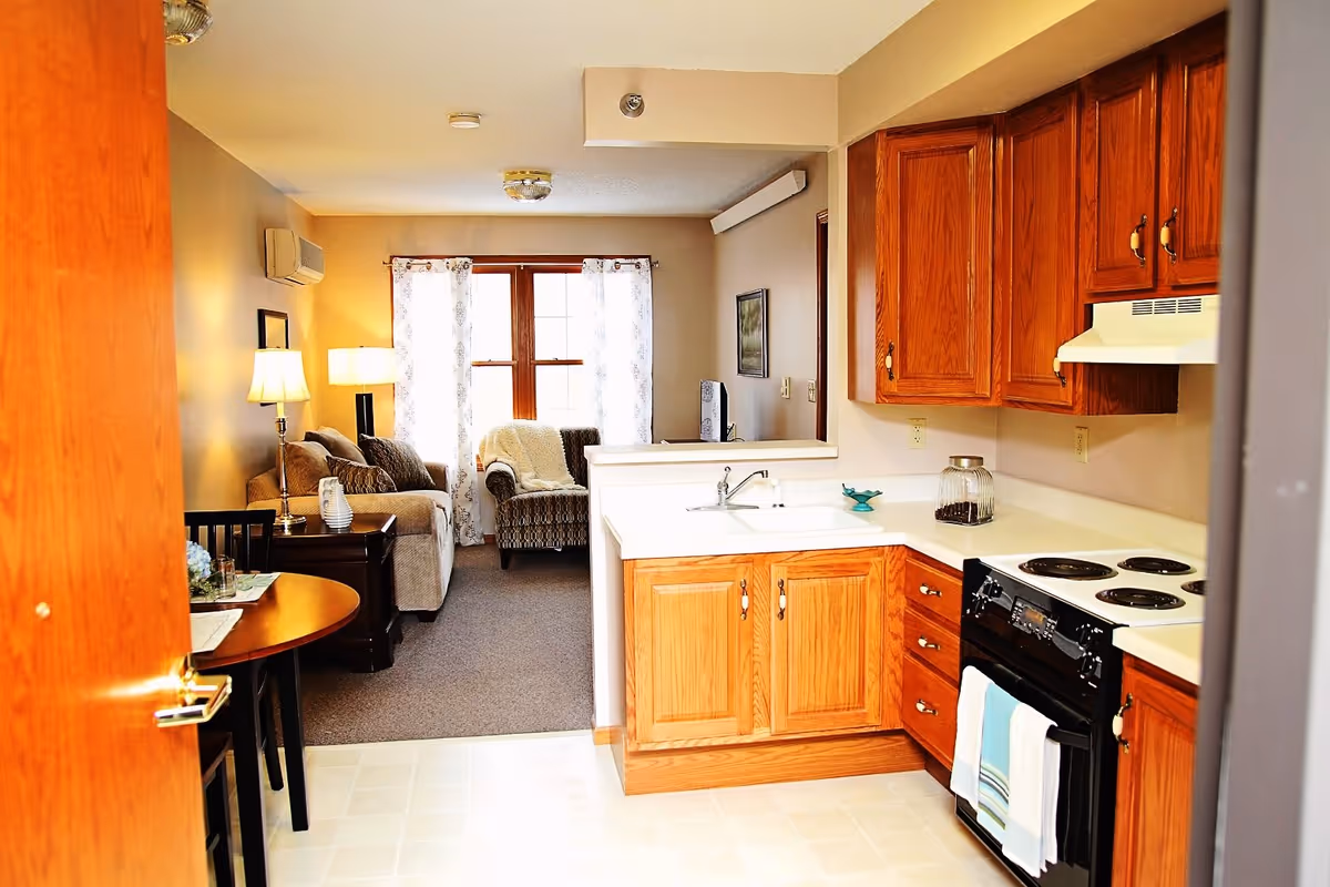 View of a cozy living area and kitchen in a senior living facility. The kitchen features wooden cabinets, a white countertop, a sink, and a black stove with two towels hanging on the handle. The living area has a beige sofa, an armchair with a white throw blanket, a side table with a lamp, and a window with sheer white curtains allowing natural light to enter.