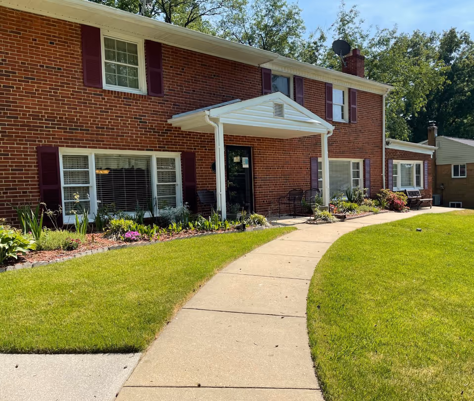 Brick senior living facility entrance with curved walkway, lawn, and outdoor seating.
