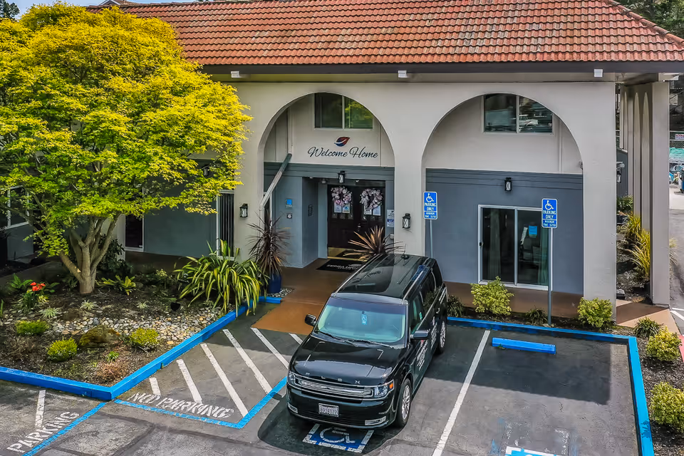Front entrance of Serra Highlands Senior Living with arched portico, landscaped planter, handicap parking spaces and a black van parked out front.