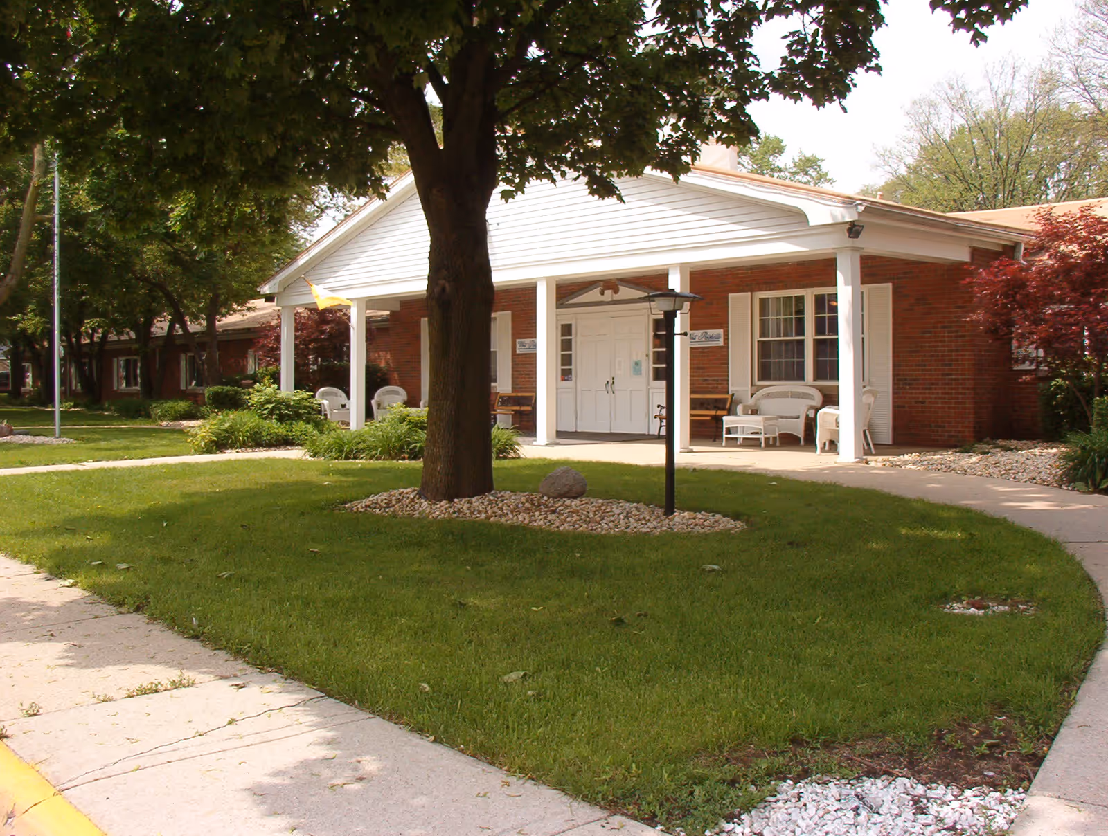 Front entrance of a single-story brick senior living facility with a covered porch, white columns, chairs, and a large tree on a green lawn.