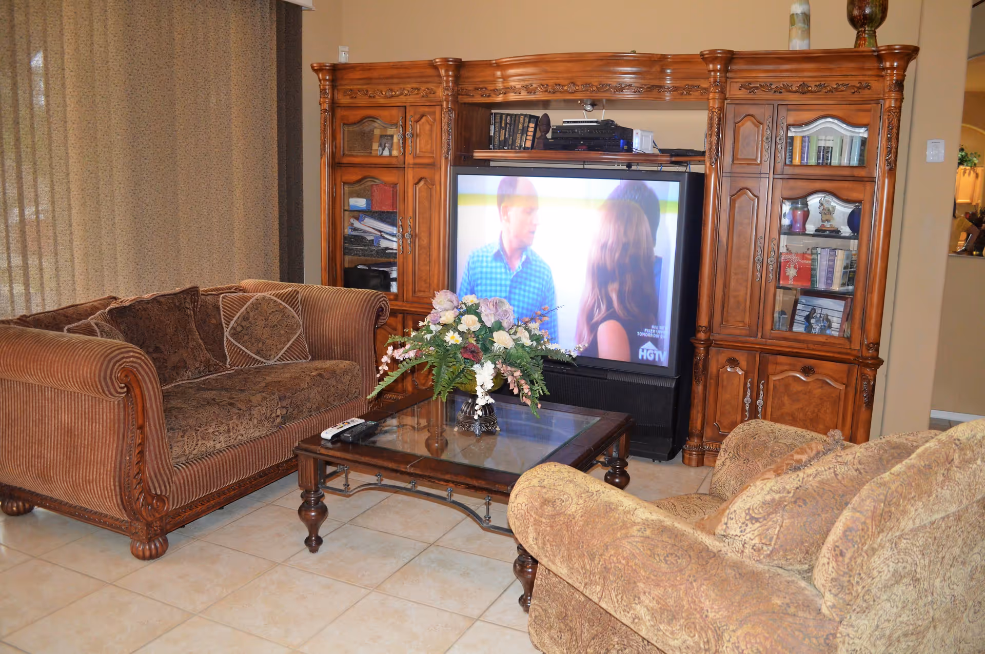 Cozy living room with patterned sofas, a glass coffee table topped with a floral arrangement, and a large wooden entertainment center with a television.