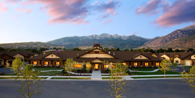 Front exterior view of River Meadows Senior Living facility at dusk with warm lights glowing from windows, surrounded by landscaped greenery and trees, with mountains in the background under a partly cloudy sky.