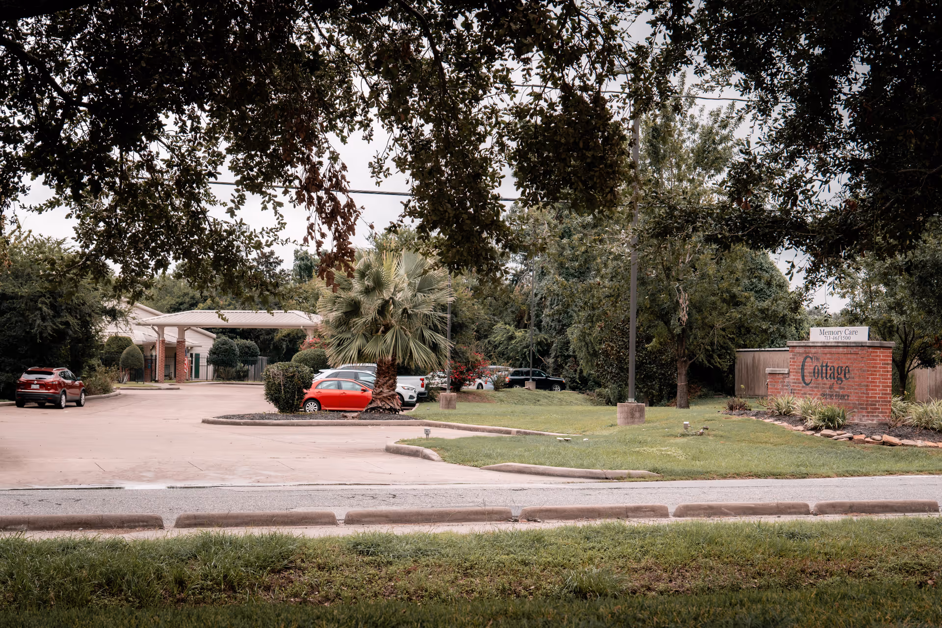 Exterior view of The Cottage of Spring Branch showing the driveway with parked cars, palm tree landscaping, and a brick entrance sign.