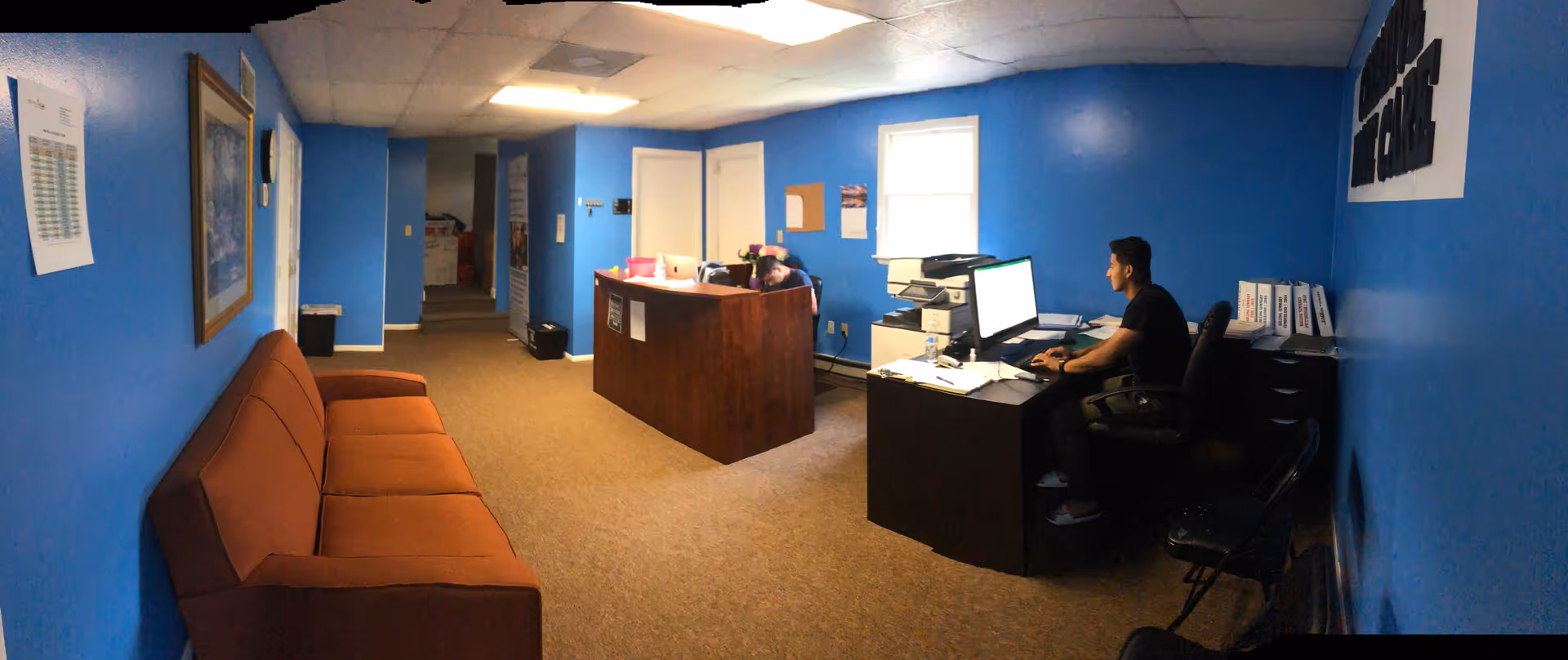 Blue-walled reception area with brown couch, desk, and staff member at a computer.