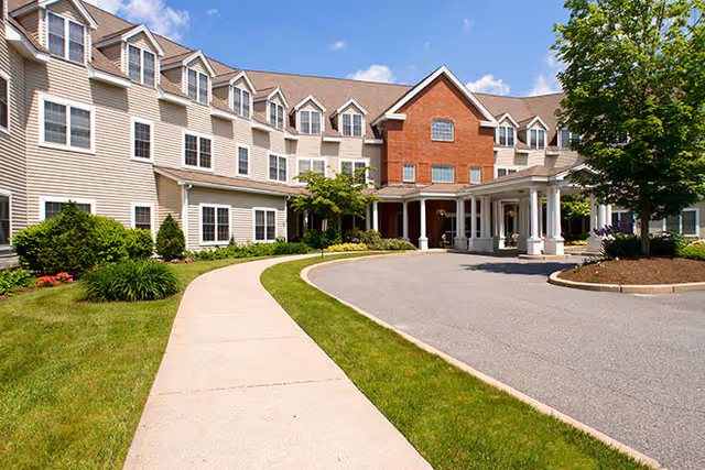 Front exterior of a multi-story senior living building with a covered entrance, curved driveway, and landscaped lawn.